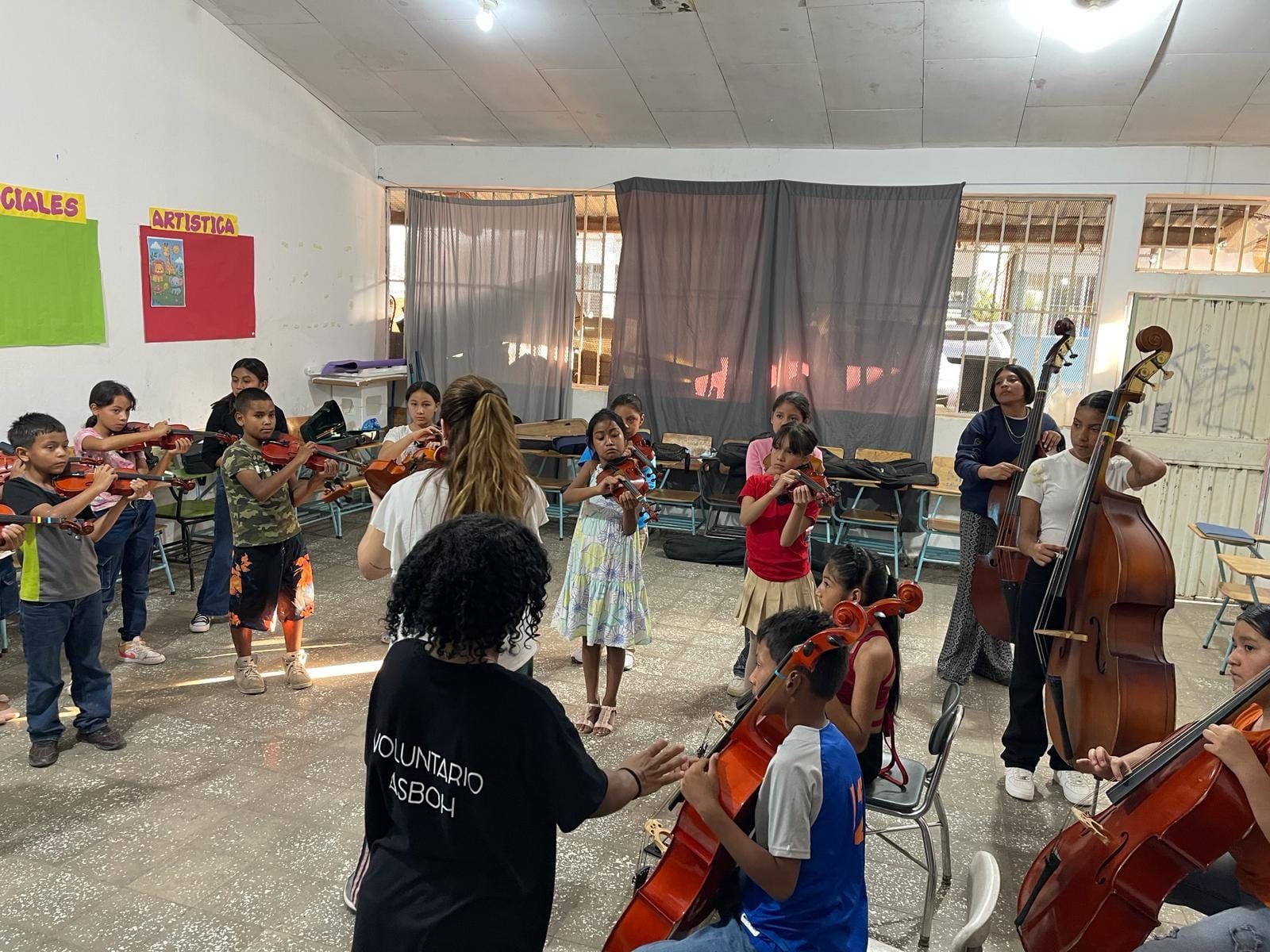 Clase de orquesta en un aula: niñas y niños tocan violines y violonchelos junto a personas voluntarias con camiseta de ASBOH