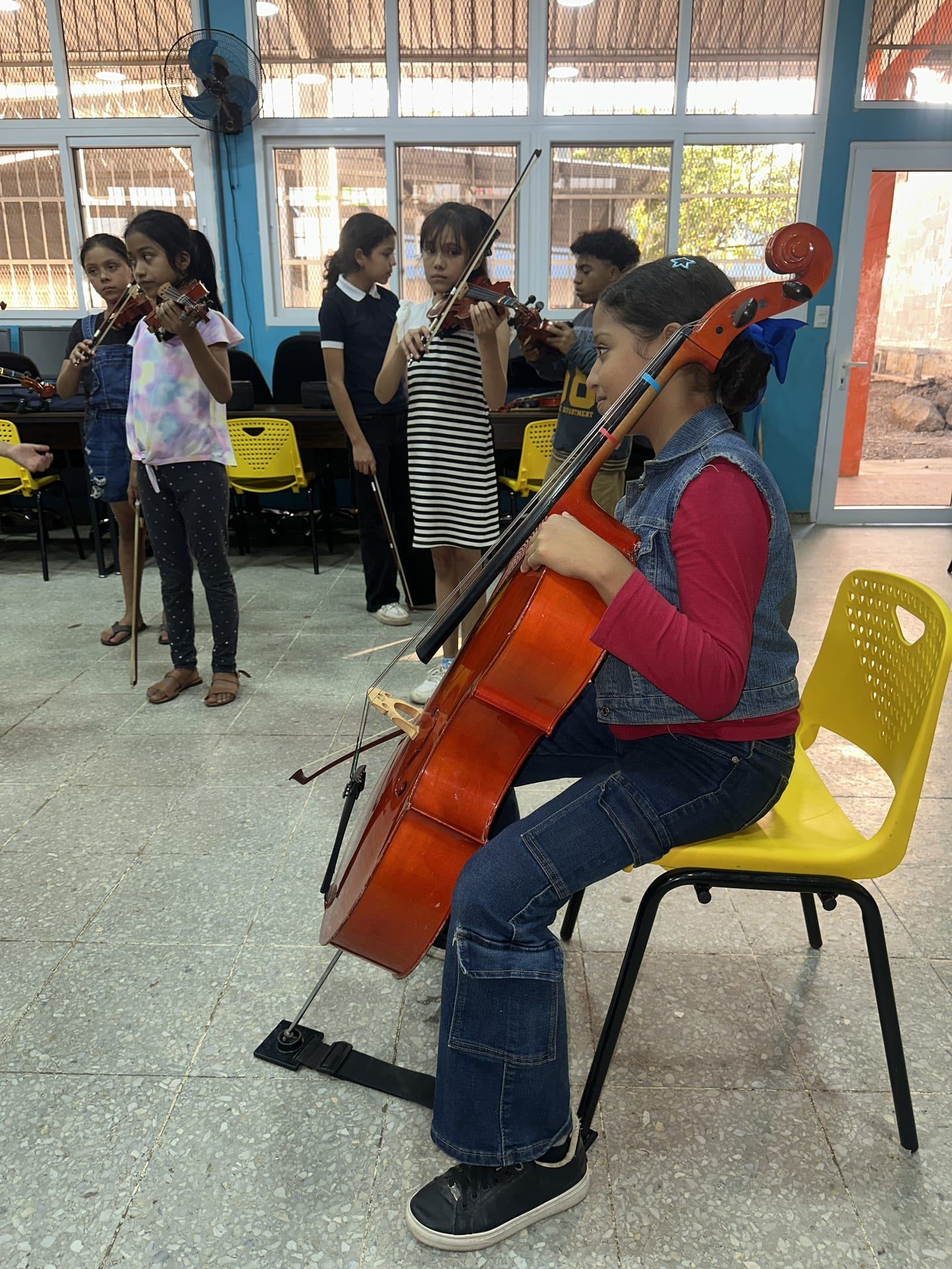 Una niña toca el violonchelo sentada en una silla amarilla, con otras niñas con violines detrás