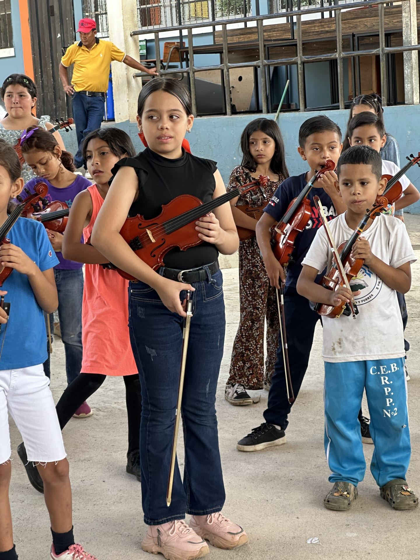 Niñas y niños con sus violines y arcos, atentos a la clase en el patio del centro educativo