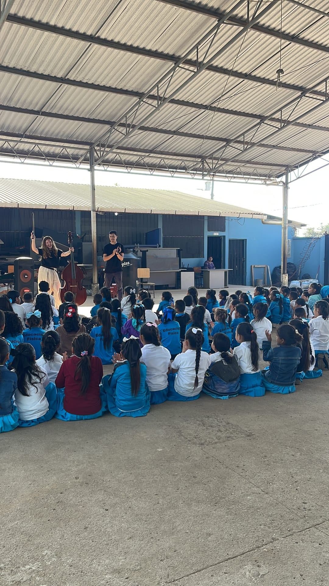 Niñas y niños sentados en el piso de un patio techado miran una presentación musical