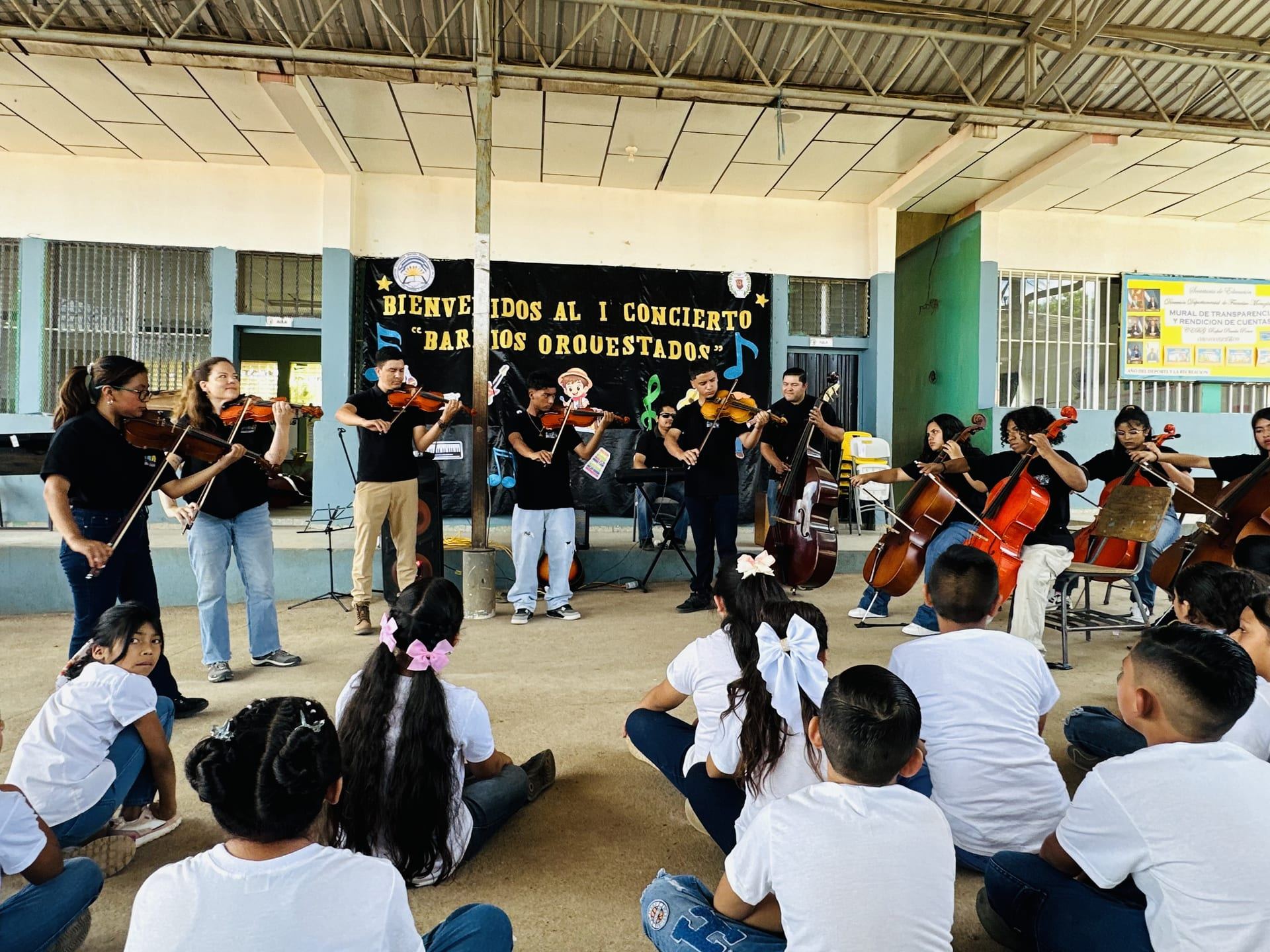 La orquesta de niñas, niños y jóvenes toca bajo el cartel del concierto, ante sus compañeros sentados en el piso