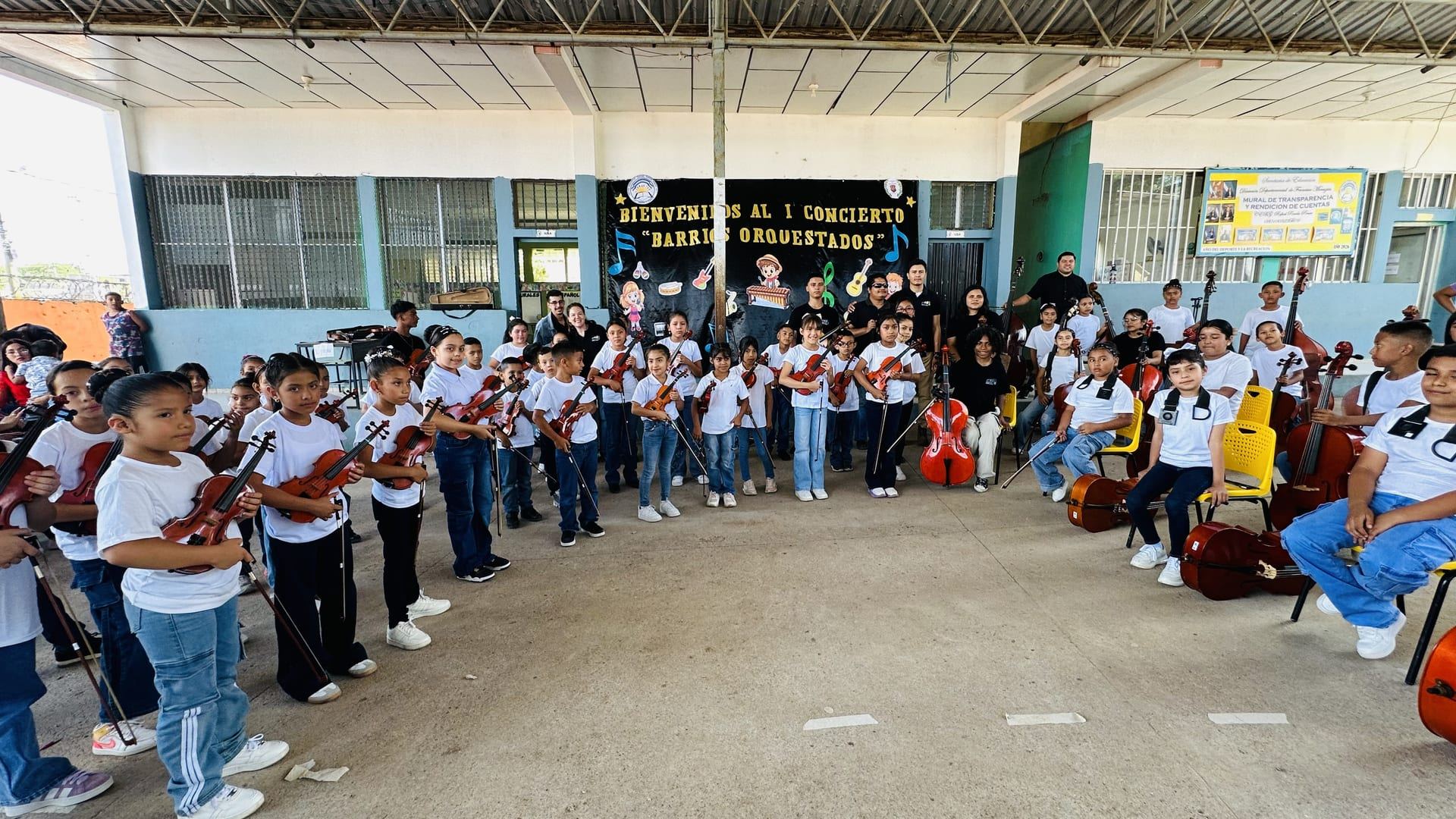 Panorámica del concierto del 18 de junio de 2026: alumnos con violines y violonchelos forman un semicírculo frente a un cartel de bienvenida