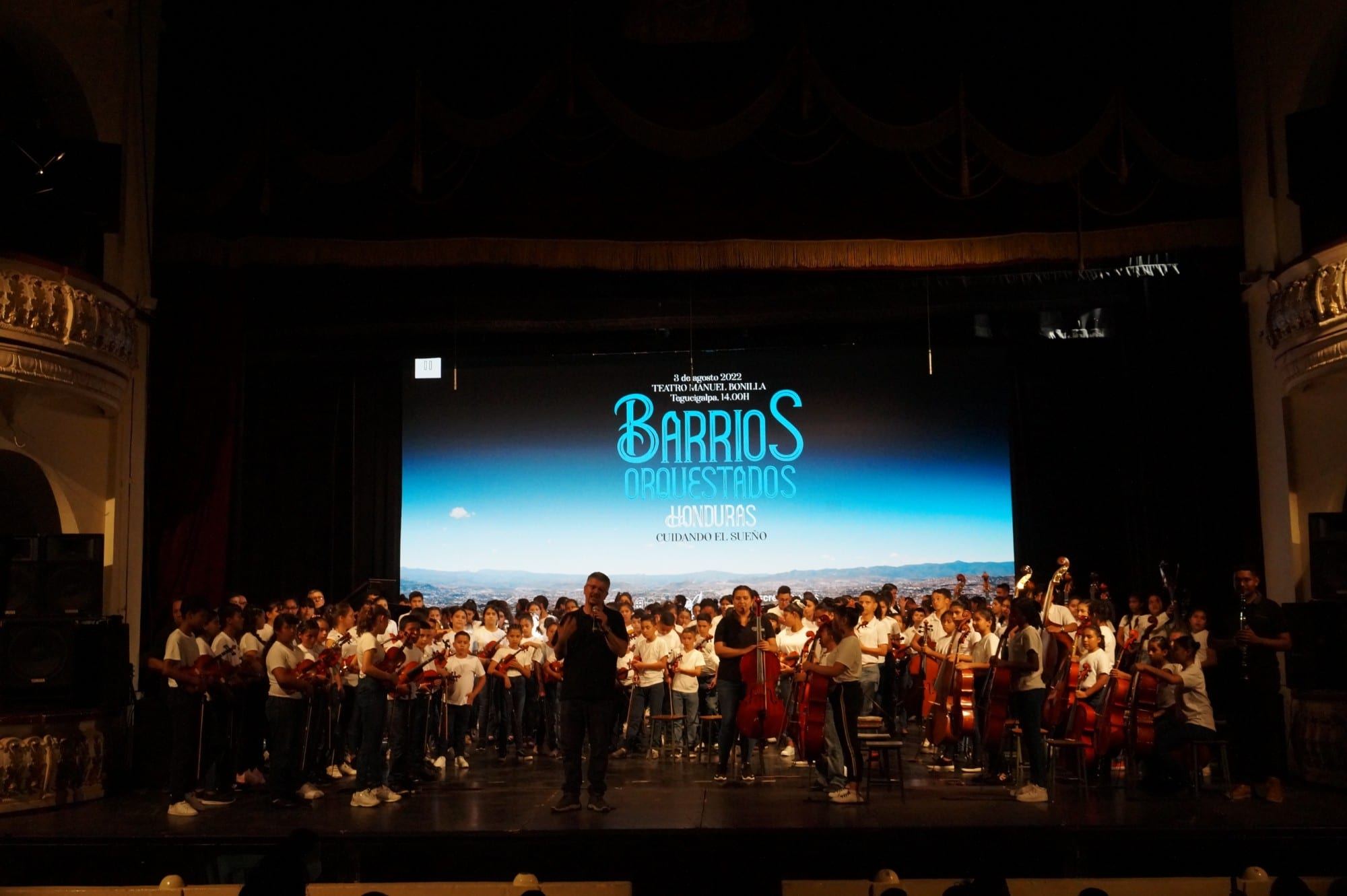 Escenario del Teatro Manuel Bonilla, en Tegucigalpa, lleno de niñas, niños y jóvenes con sus instrumentos frente a una pantalla que dice «Barrios Orquestados Honduras. Cuidando el sueño»