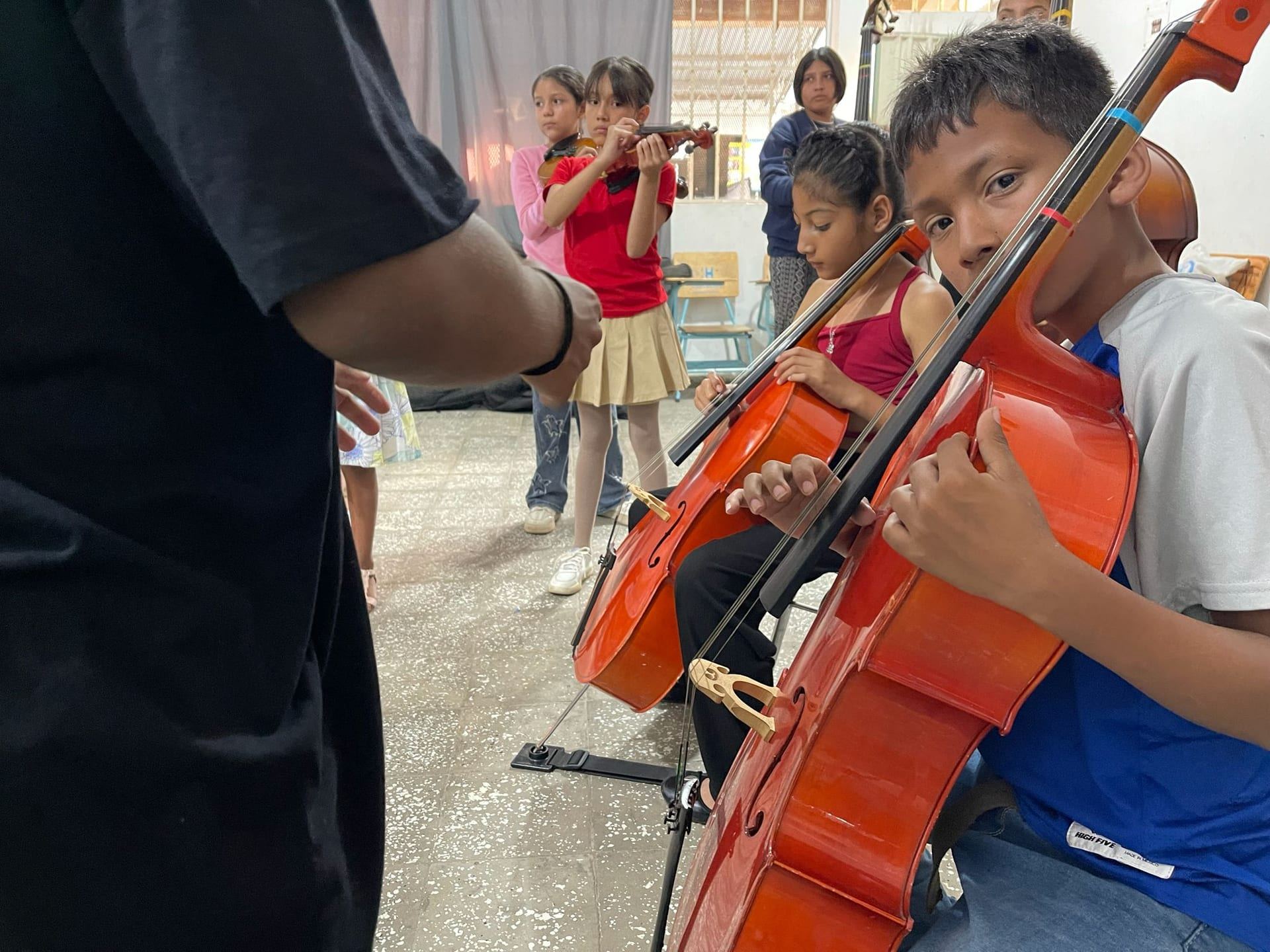 Un niño toca el violonchelo en primer plano durante una clase; al fondo, otros alumnos