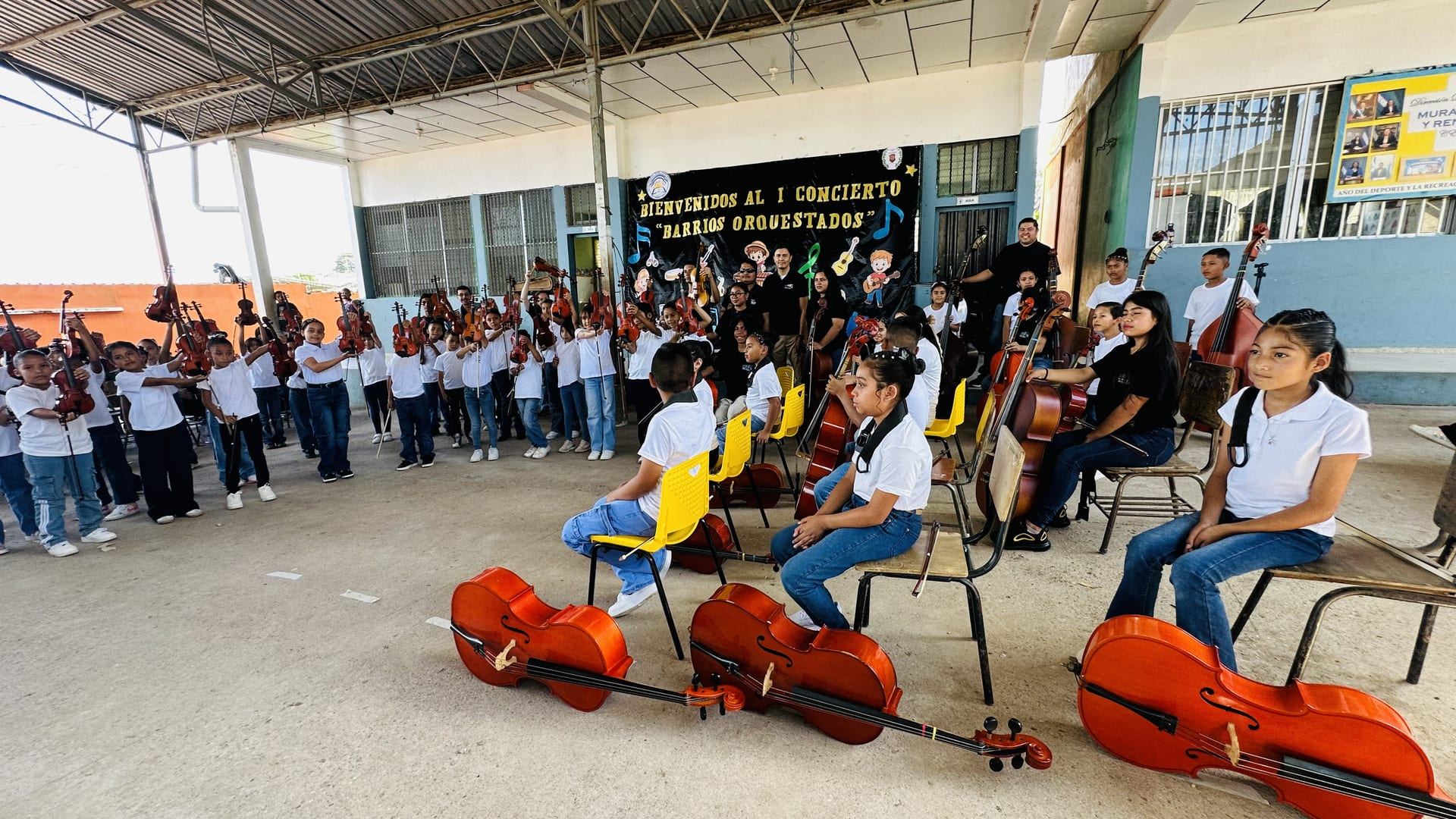 Los alumnos con violines y violonchelos esperan en semicírculo; en primer plano, tres violonchelos en el piso